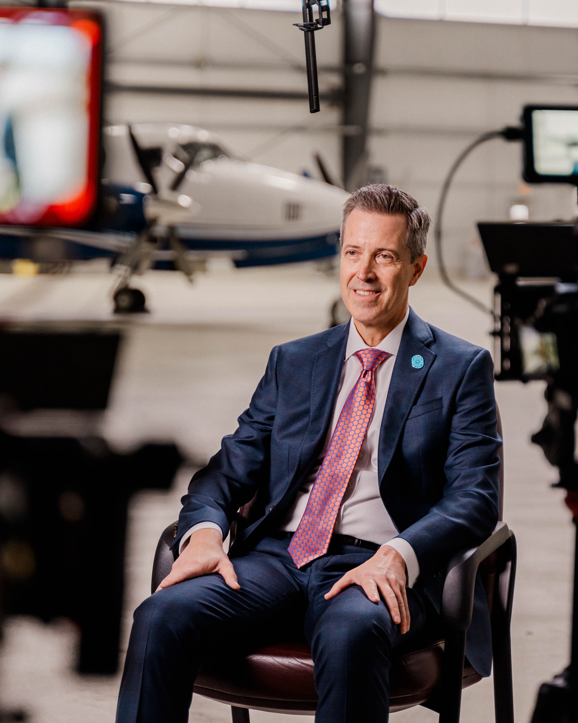 a man smiles in front of storyteller studios film cameras under lights in an airplane hanger wearing a navy suit and plum colored tie