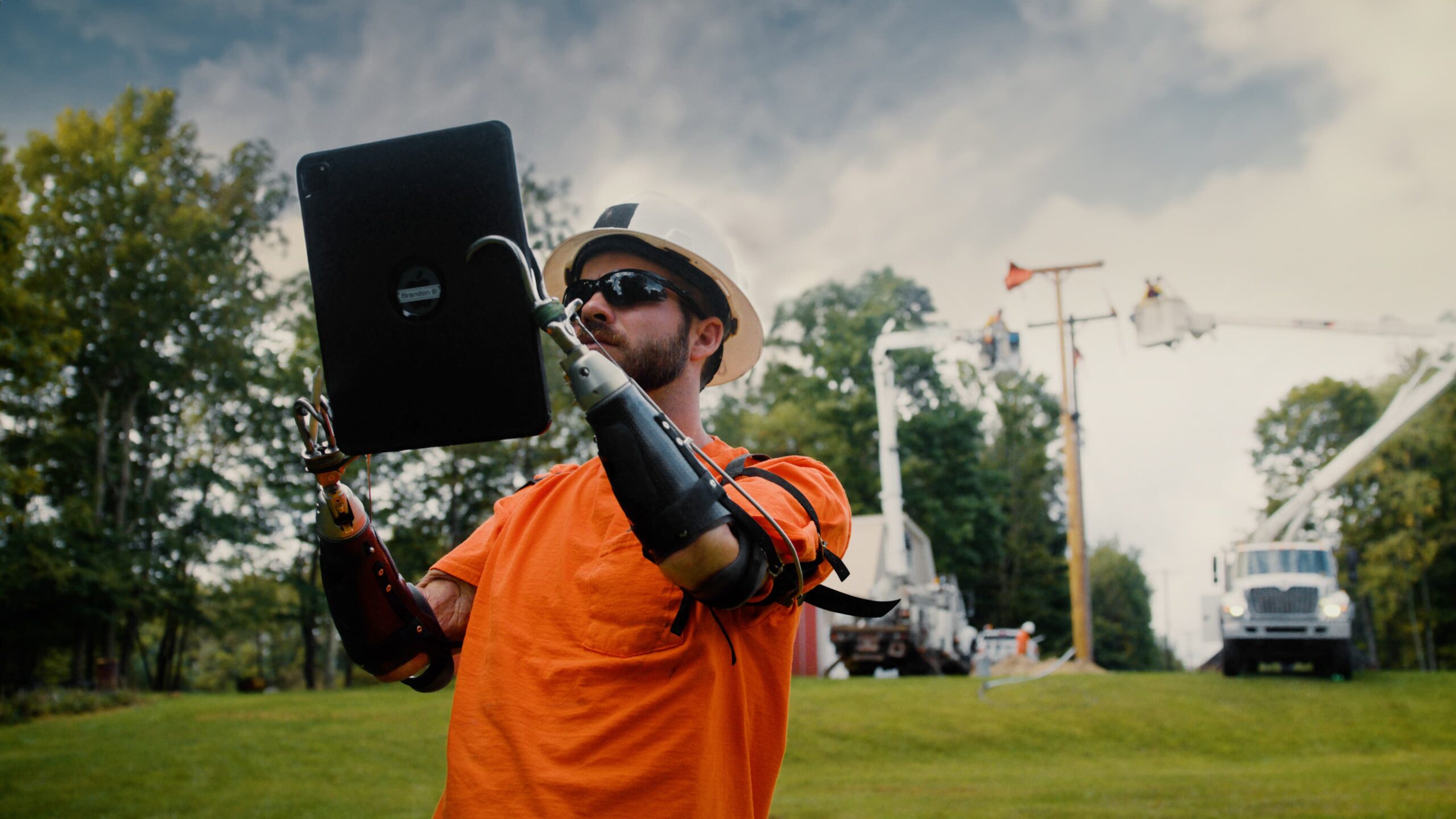 a lineman wearning an orange shirt and white hard hat checks wire schematics on an ipad in the field