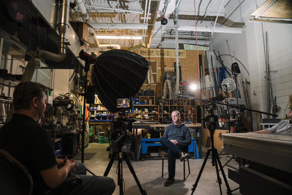 A behind-the-scenes view of a professional video interview setup inside a workshop. A man in a dark hoodie sits for the interview, surrounded by tools, shelves, and industrial equipment. The production team has set up a large softbox light, multiple cameras, and a boom microphone to capture high-quality footage. The environment features exposed pipes and a high ceiling, creating a raw, authentic backdrop.