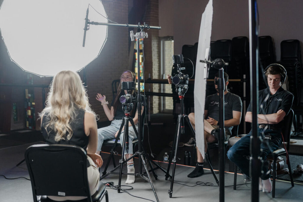 A behind-the-scenes look at a professional video interview setup, featuring a seated interviewee illuminated by a large softbox light. A camera operator and production team members are visible in the background, adjusting equipment and monitoring audio. The setting appears to be an indoor venue with stacked chairs and stained-glass windows in the background.