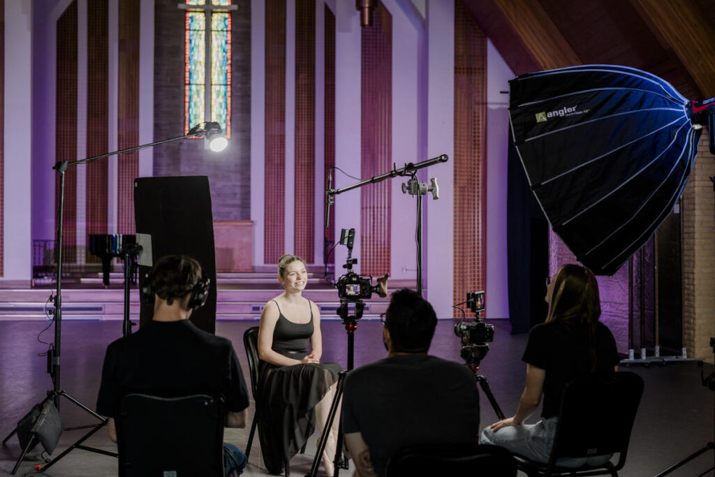 A behind-the-scenes look at a professional video interview setup inside a grand venue with stained glass windows and high ceilings. A young woman in a black dress sits in the interview chair, gesturing as she speaks. A large softbox light, boom microphone, and cameras surround her, while the production crew monitors the setup. The background features an elevated stage with warm ambient lighting.