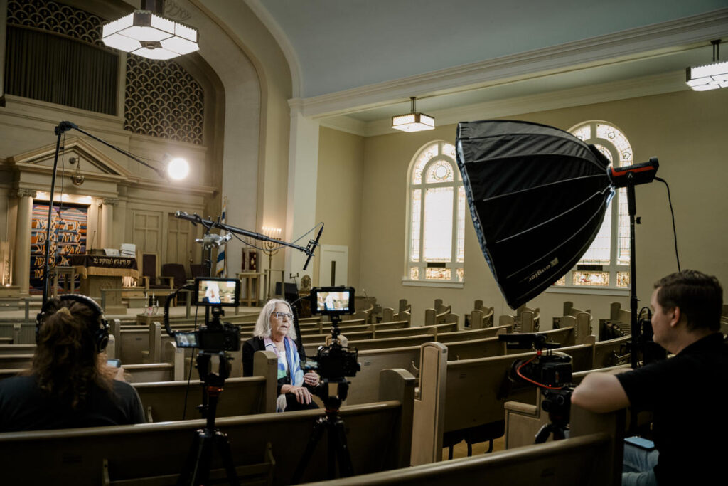 A professional video interview setup inside a synagogue, featuring a woman seated in a pew while being filmed by a production team. The setup includes multiple cameras, a boom microphone, and a large softbox light for controlled lighting. The historic architecture and stained-glass windows in the background add depth to the scene.
