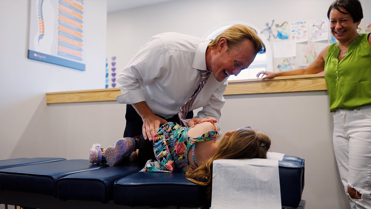 Dr. Bryne Willey of AlignLife smiles warmly as he gently adjusts a young girl named Ginger on a chiropractic table. Ginger, wearing a colorful dress, laughs as she interacts with Dr. Willey. In the background, a woman watches with a joyful expression, standing in a bright, welcoming clinic space decorated with children’s artwork and a spinal health poster.