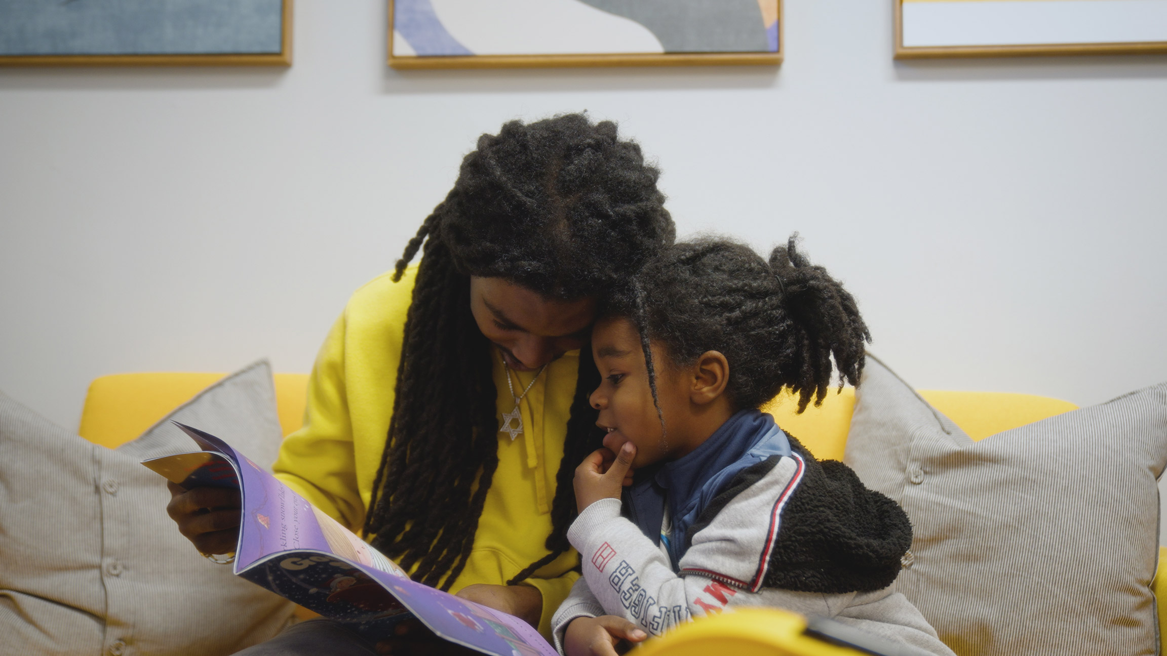 A father and young son sit close together on a yellow couch, reading a colorful children’s book. Their foreheads touch as they share a quiet moment.
