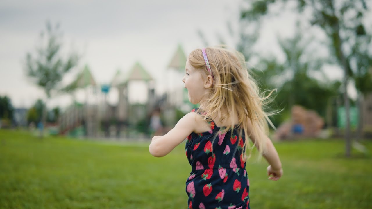A young girl with blonde hair runs through a grassy park toward a playground, wearing a dress with red strawberries. Her face is joyful and free.