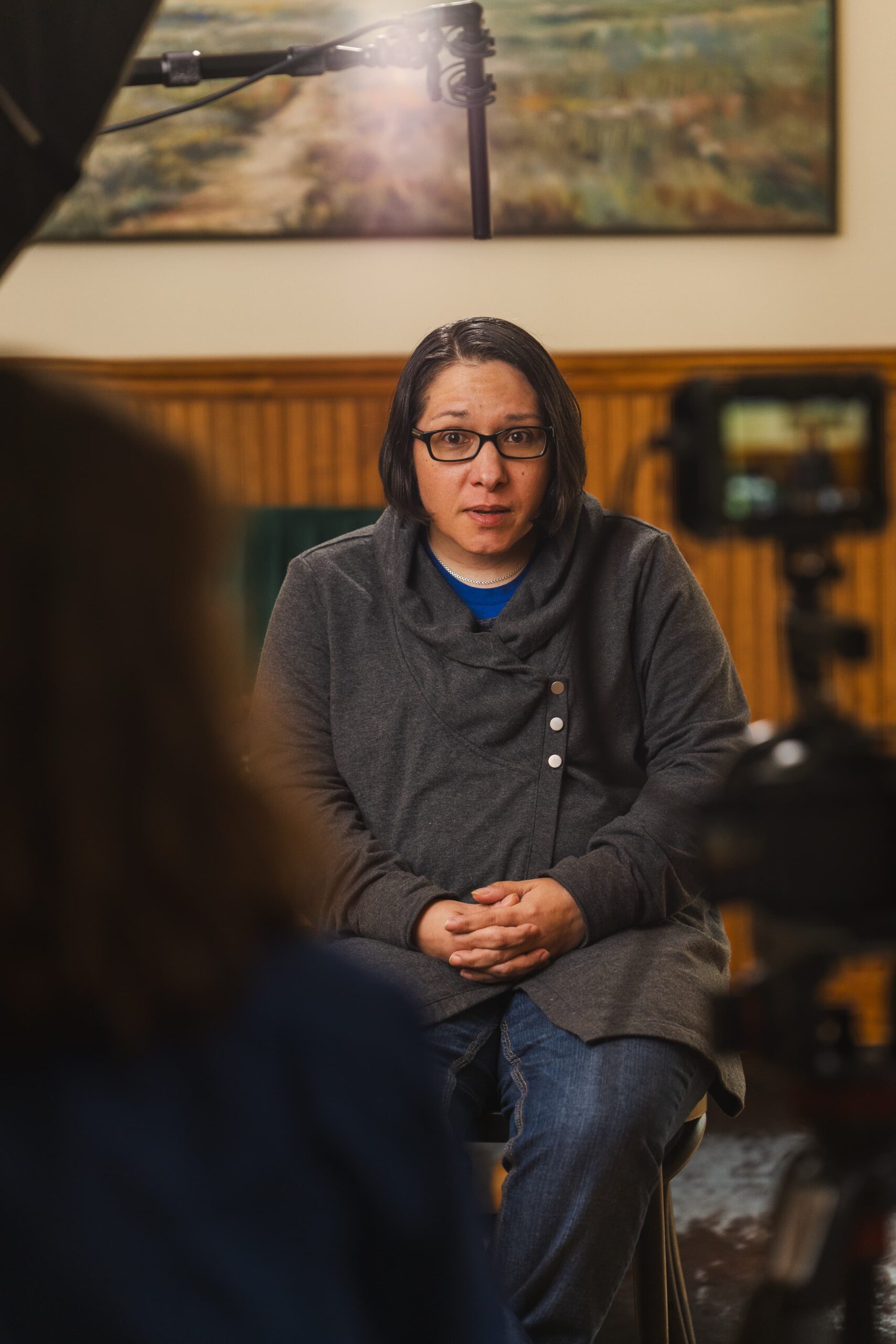 A person wearing glasses, a dark gray sweater, and jeans sits for an on-camera interview, hands folded, speaking directly toward the lens. Professional lights and a boom microphone are visible above, with a camera capturing the scene in the foreground.