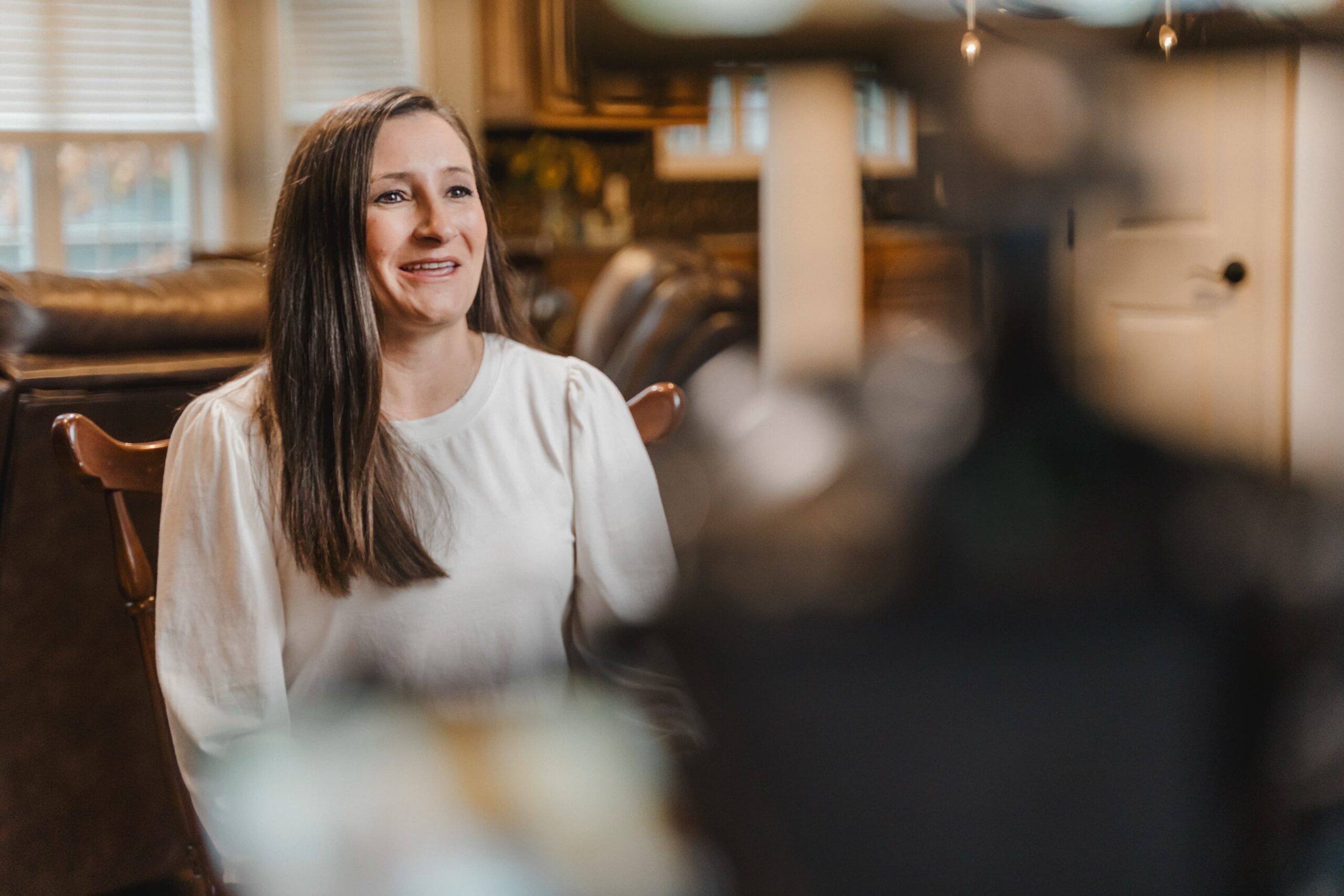 A woman with long brown hair and a white blouse sits in a warmly lit living room, smiling during a filmed interview. A camera and lights are partially visible in the blurred foreground.