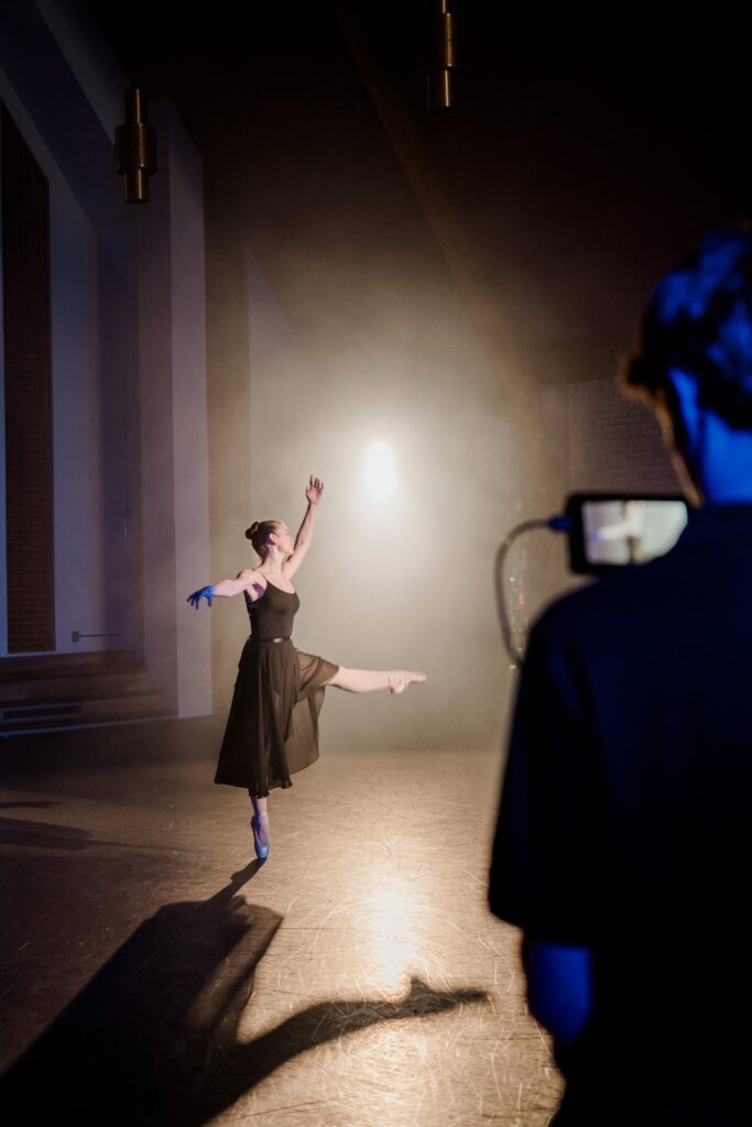 Professional video production setup filming ballet dancer in flowing brown dress performing graceful pose in dramatic studio lighting