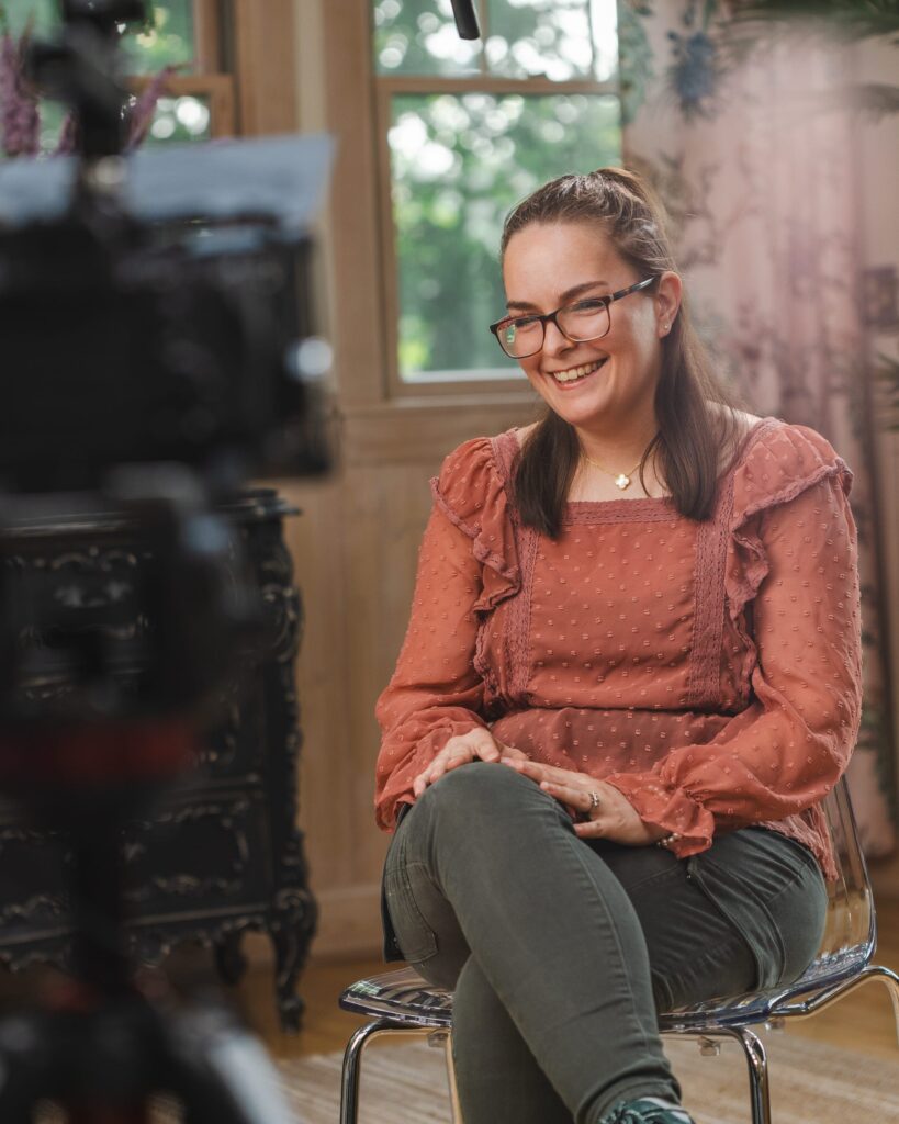 A young woman sits smiling during an on-camera interview, wearing glasses, a textured rust-colored blouse, and green pants. She is seated on a clear chair in front of a softly lit window, with a video camera and microphone visible in the foreground.