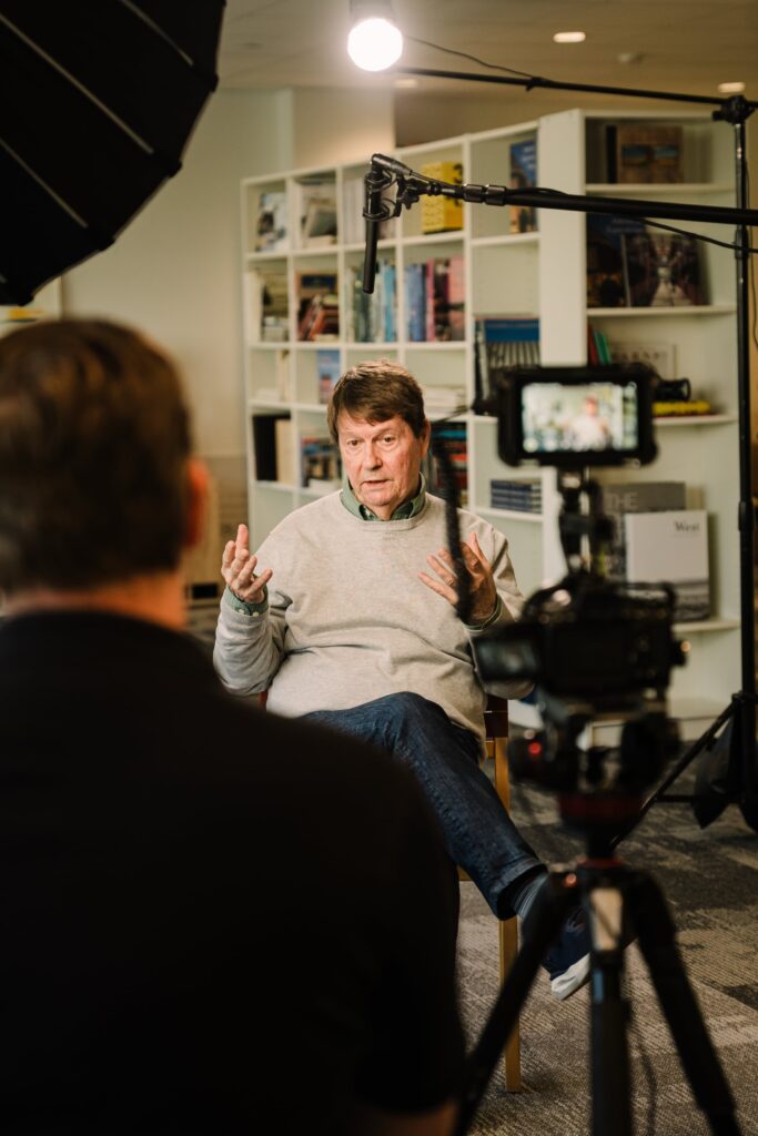 John Blackburn, founder of Blackburn Architects, being interviewed on camera by Storyteller Studios with professional lighting and boom microphone in his office with bookshelves behind him.