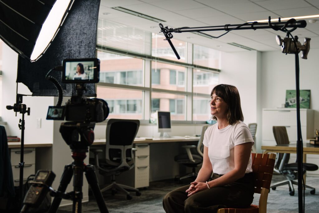 Elizabeth Charpentier, Project Manager at Blackburn Architects, being filmed during brand video interview in modern office conference room. Professional cinema camera on tripod with boom microphone and lighting setup visible.