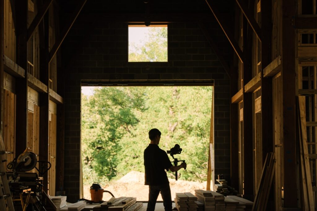 Storyteller Studios videographer silhouetted in doorway of Blackburn Architects construction site holding professional camera equipment. Natural lighting streaming through barn opening with wooden framework visible.