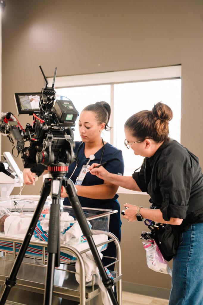 Hair and makeup artist prepares an actor playing a nurse beside a newborn on the set of the IDPH hearing screening PSA
