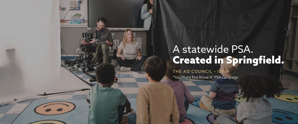 Behind the scenes in a Springfield classroom: Storyteller Studios producer Chris Costello operates camera while Lydia Stuemke directs children for the Illinois Department of Public Health’s “You Might Not Know It” PSA campaign. Text over image: “A statewide PSA. Created in Springfield.” The Ad Council, IDPH.