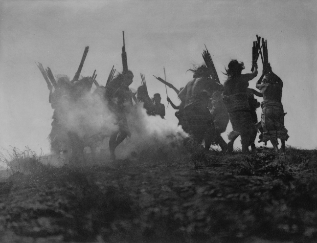 Several Kwakiutl people dancing in a circle around a smoking fire, in an effort to cause a sky creature, which they believe swallowed the moon, to sneeze thereby disgorging it. Photograph by Edward S. Curtis, 1914.