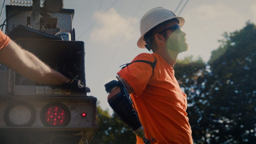 a lineman in a bright orange shirt and white hard hat leans against a utility truck to rest