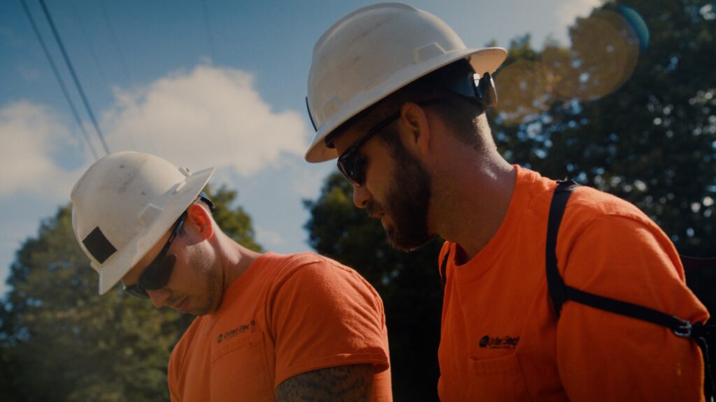 two lineman wearing bright orange shirts and white hard hats work outside on a sunny day